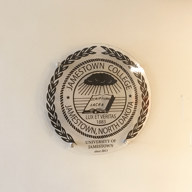 Round emblem with 'Jamestown College' text and design on a beige background on campus in Jamestown, ND.